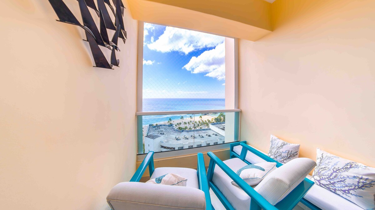 A serene balcony features comfortable seating, allowing for relaxation while enjoying unobstructed views of the beach and ocean. Natural light fills the space, with decorative elements complementing the coastal ambiance. Shutters provide a blend of privacy and openness to the picturesque surroundings.