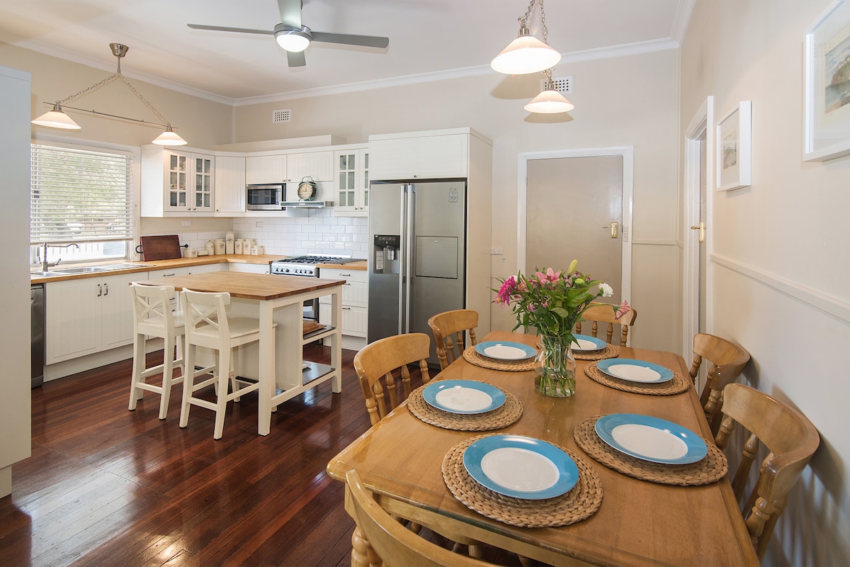 A modern kitchen features a central island with bar seating, complemented by a dining area with a wooden table and woven placemats. Ample cabinetry and appliances are visible, while natural light streams through a nearby window, illuminating the polished wooden flooring.