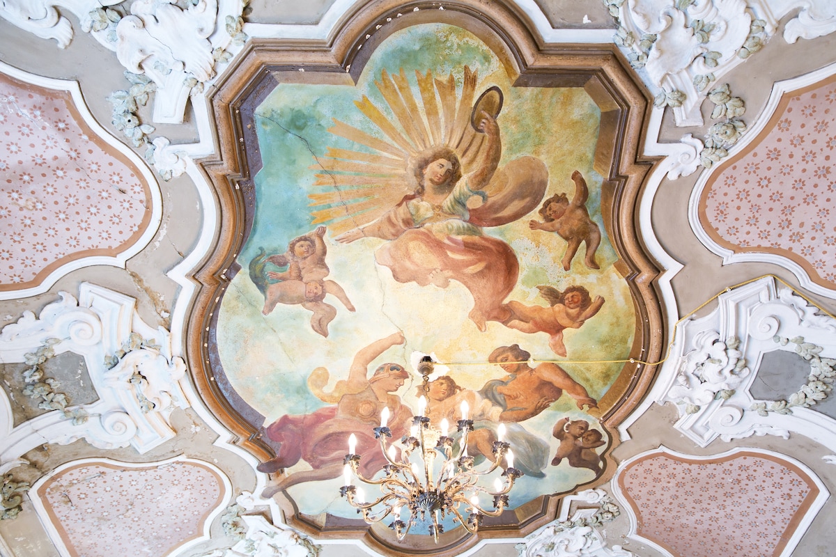 An intricately painted ceiling features a vibrant fresco with cherubs and celestial figures, surrounded by ornamental plasterwork. A chandelier hangs gracefully from the center, illuminating the artistic details. The soft color palette showcases the historic artistry typical of the Sicilian rococo style.