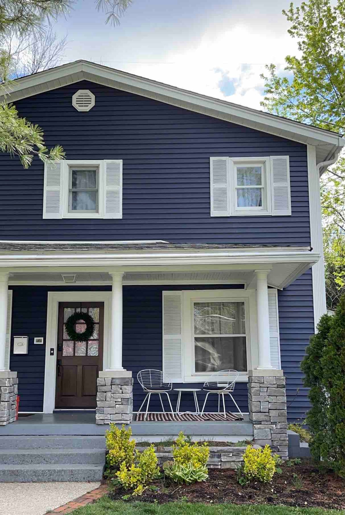 The exterior of a recently remodeled two-story home is highlighted by blue siding and white trim. A welcoming front porch features two white chairs and a small table, framed by well-manicured shrubs and greenery. A decorative wreath adorns the front door.