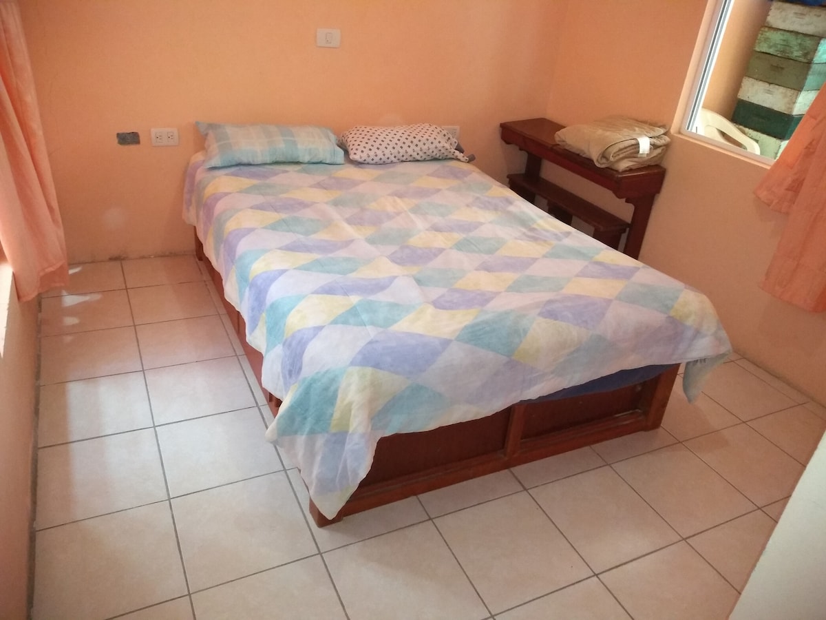 The room features a spacious bed with a colorful patterned comforter, flanked by two pillows. Soft orange curtains frame a window, allowing natural light to enhance the peach-colored walls. A small table is present beside the bed, contributing to the room's practicality.