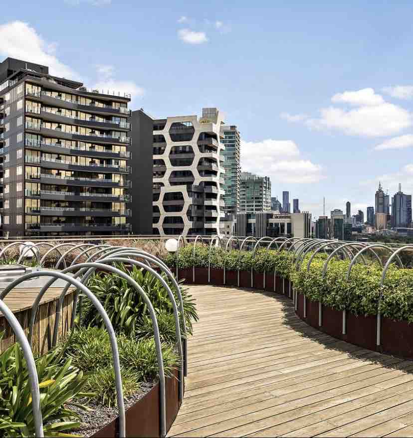 A rooftop garden pathway is lined with greenery and modern buildings in the background. The walkway features a wooden surface, and curved metal arches frame the vibrant plant life. The city skyline is visible on the horizon, showcasing the urban setting.