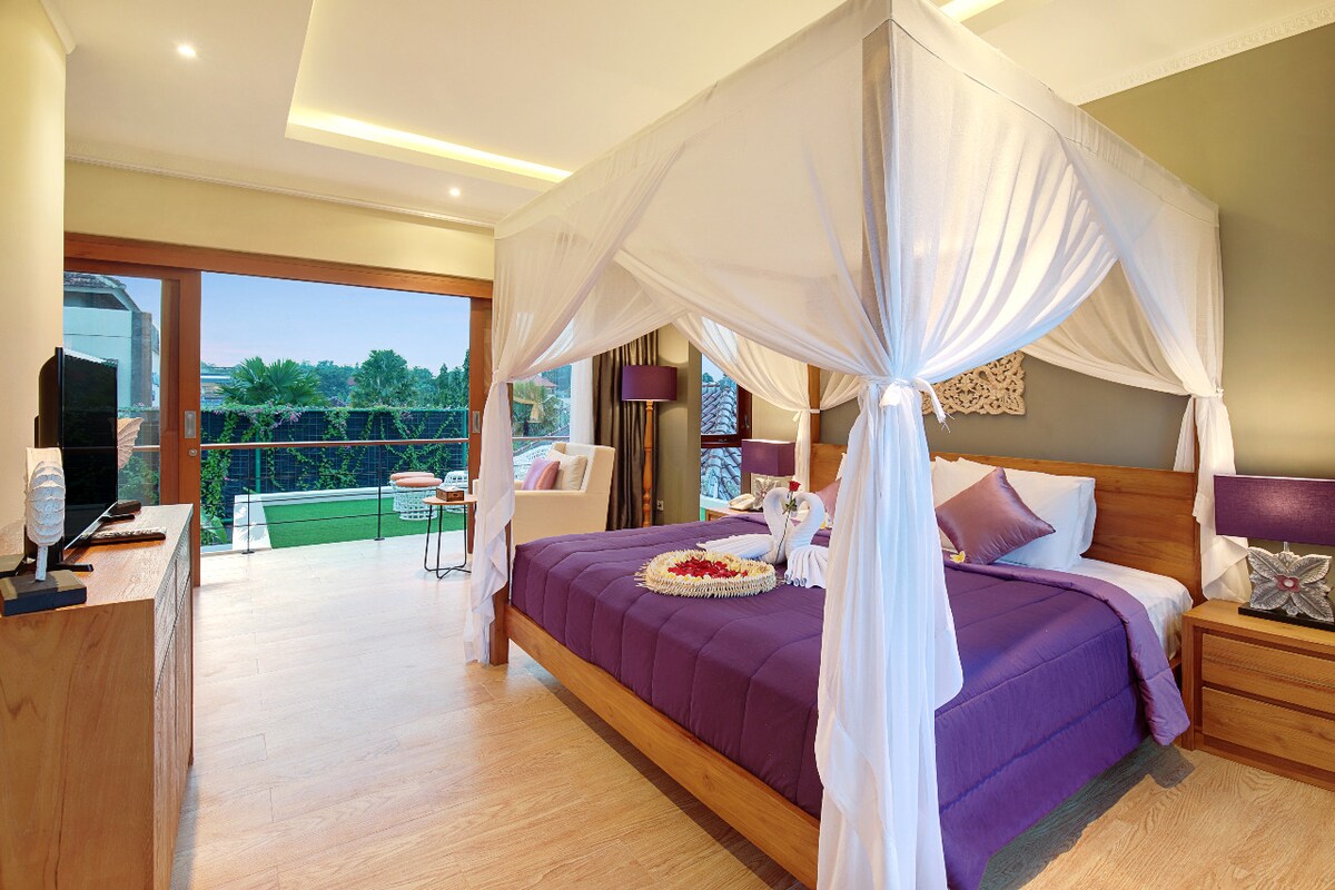 A spacious bedroom features a four-poster bed draped with white fabric, complemented by purple bedding. Large glass doors open to a private terrace with views of greenery, while natural light brightens the room. A minimalist wooden dresser holds a flat-screen TV and decorative items.