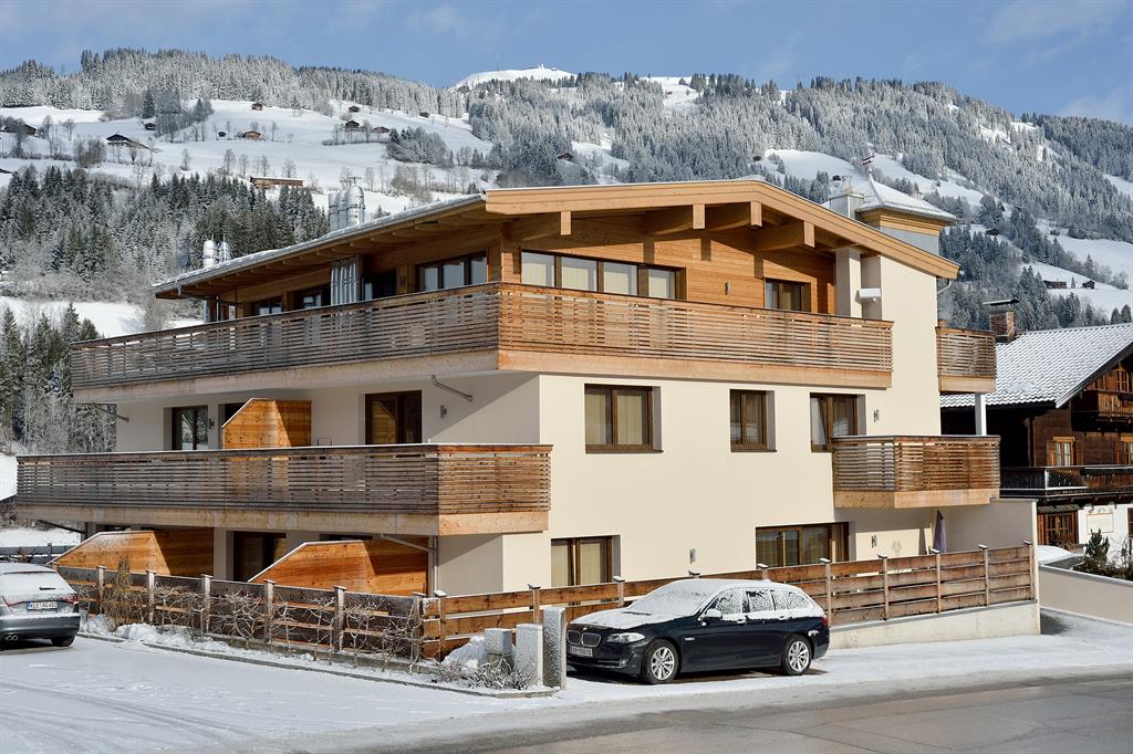 The building features a modern architectural design with multiple balconies and wooden accents. Snow covers the surrounding landscape, while distant hills are lined with trees dusted in white. A parking area with vehicles is visible in the foreground.