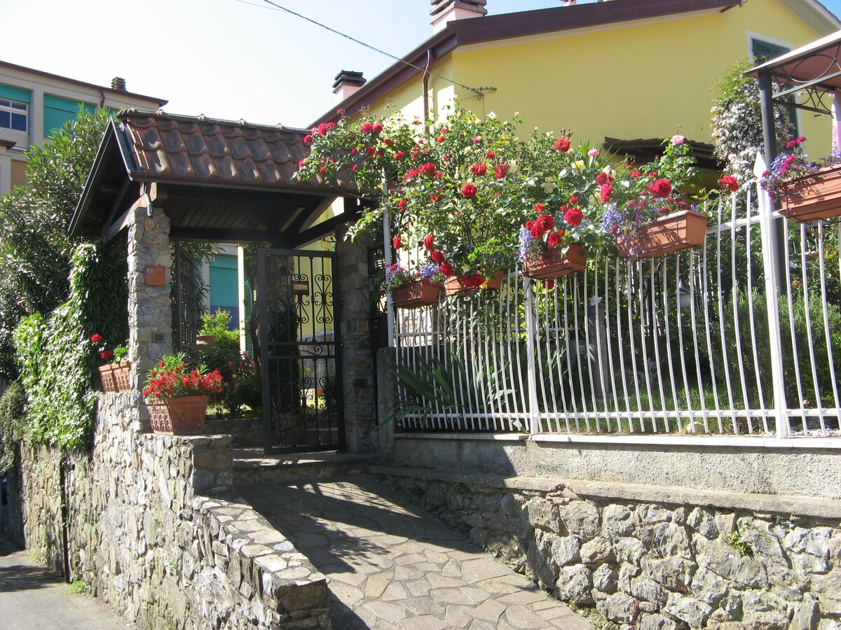 A charming entrance is framed by a stone wall and a garden filled with blooming flowers. A pathway leads to a gated entrance, while flower pots and railing planters add vibrant color to the scene, enhancing the inviting ambiance of the property.