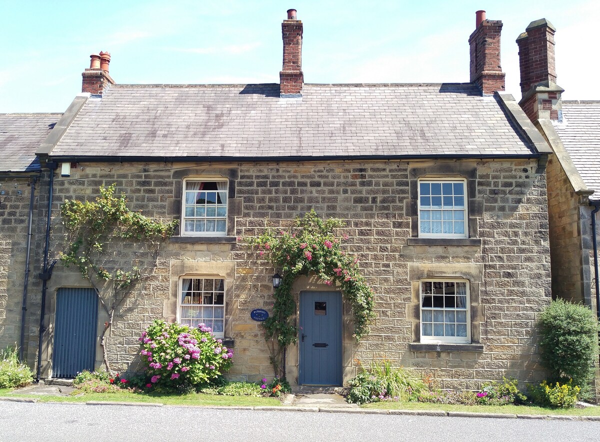 The stone façade of the cottage showcases its historic charm, with climbing plants and colorful flowers adorning the entrance. Window panes reflect natural light, while the slate roof and chimney pots add character. A welcoming door is centrally located, enhancing the inviting appearance.