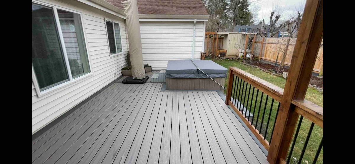 The deck is visible, showcasing a hot tub covered with a protective lid. A railing borders the deck, and a large umbrella is positioned to provide shade. The backyard features a well-maintained lawn, enhancing the outdoor space.