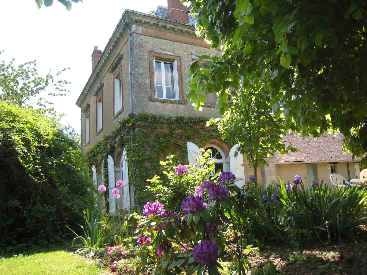 The exterior of a charming house is surrounded by lush greenery and vibrant flowers, including pink rhododendrons. White shutters frame the windows, while ivy climbs the building's walls, contributing to the natural beauty of the secluded garden.