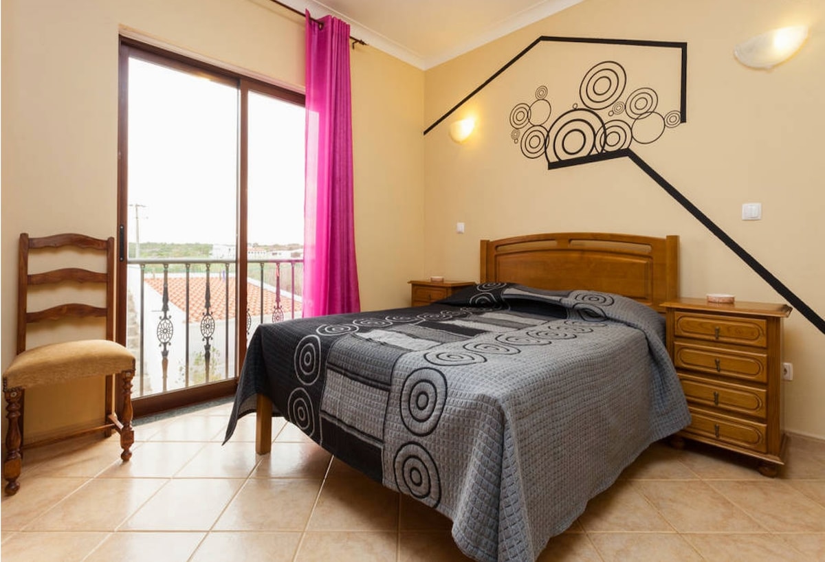 A cozy room is furnished with a double bed covered with a patterned gray blanket. A wooden nightstand and a chair are present beside the bed. Large glass doors offer natural light and open to a balcony with views, complemented by a decorative wall design.