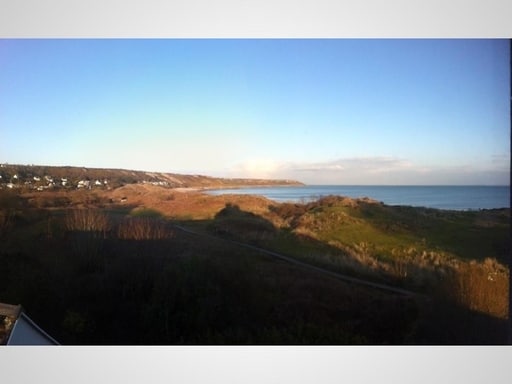 A panoramic view showcases the coastline, with stretches of sandy beach and gentle waves meeting the shore. Rolling dunes are visible in the foreground, while distant hills are faintly outlined against the clear sky. The scene conveys a sense of openness and tranquility.