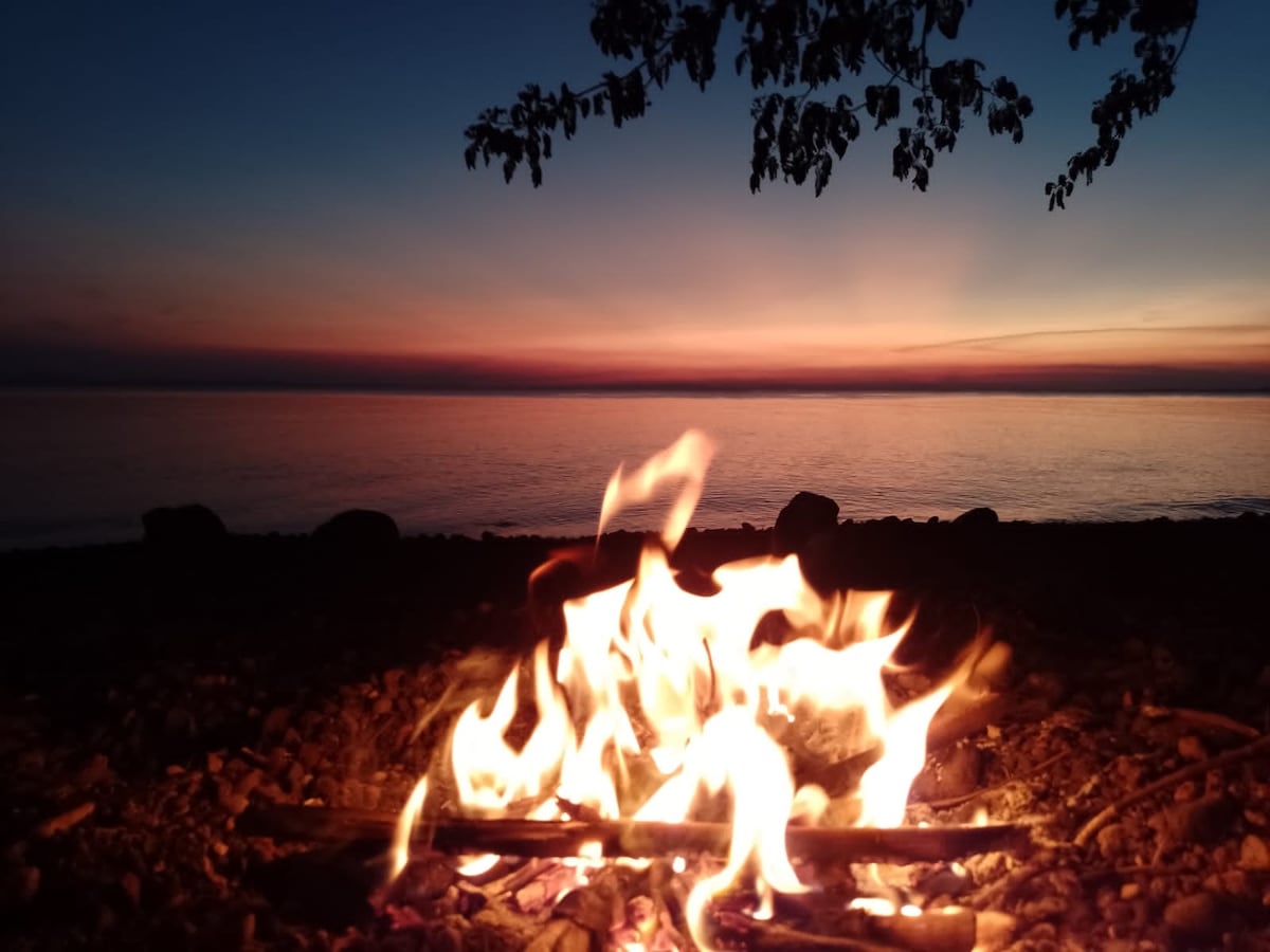 A calming bonfire flickers at the edge of a tranquil shoreline, casting a warm glow as the sun sets over the water. The horizon is marked by a gradient of colors, transitioning from soft oranges to deep blues, creating a serene backdrop.