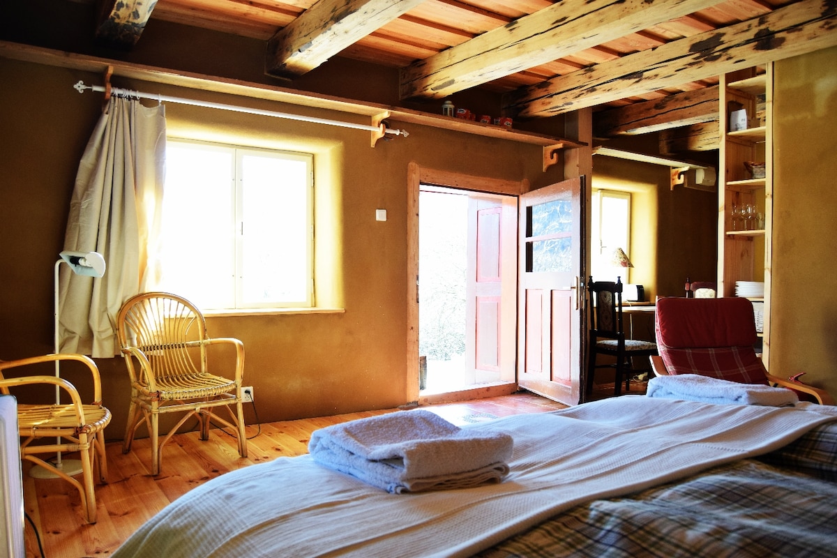 A cozy room features clay plastered walls and exposed wooden beams. Sunlight filters through a window next to a wooden door leading outside. A spacious bed with white linens is positioned in front, accompanied by a rattan chair and a red armchair, creating a welcoming atmosphere.