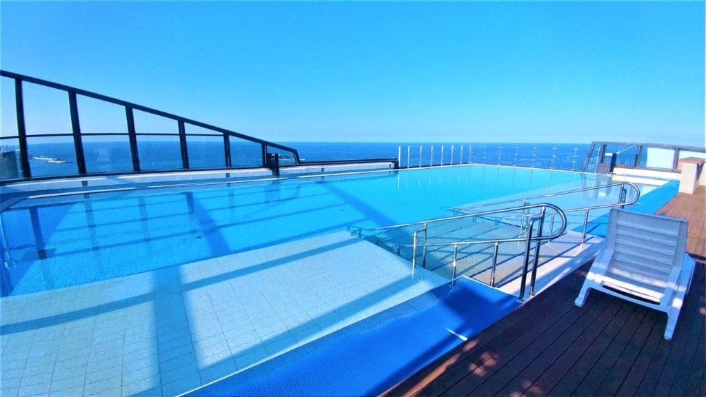 An infinity pool extends along the rooftop, offering unobstructed ocean views. The pool area is surrounded by clear glass railings and features a spacious deck with a lounge chair positioned nearby. Bright blue skies reflect in the tranquil water, creating a serene atmosphere.