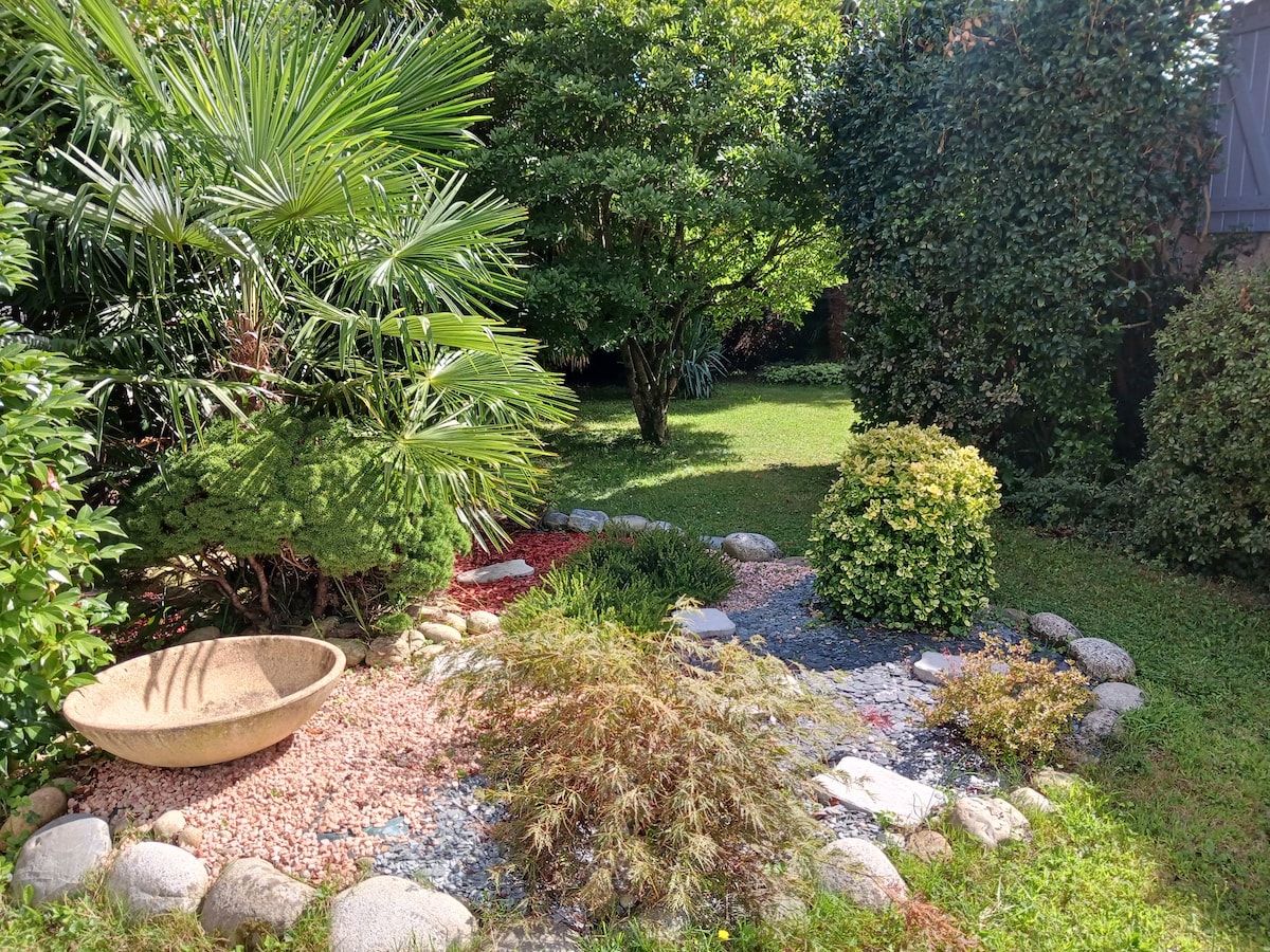 A well-maintained garden area showcases various greenery, including palm trees and shrubs. A decorative stone bowl is situated among pebbles and small rocks, leading to a lush expanse of grass surrounded by vibrant plant life.