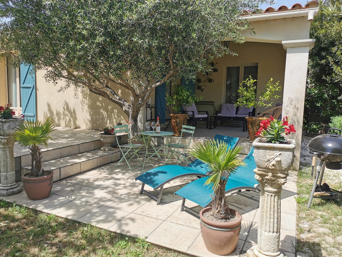 A shaded patio is adorned with lounge chairs and a dining set surrounded by potted plants. A stone pedestal features decorative elements, and a barbecue is visible. The area benefits from the shade of an olive tree, creating a tranquil outdoor space for relaxation.