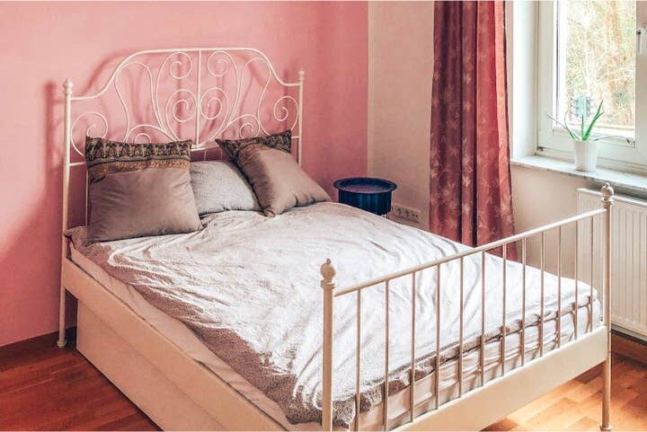 A spacious bed with decorative pillows is positioned against a soft pink wall. A window allows natural light to fill the room, while a small potted plant is visible on the windowsill. Light-colored bedding adds to the calm ambiance.