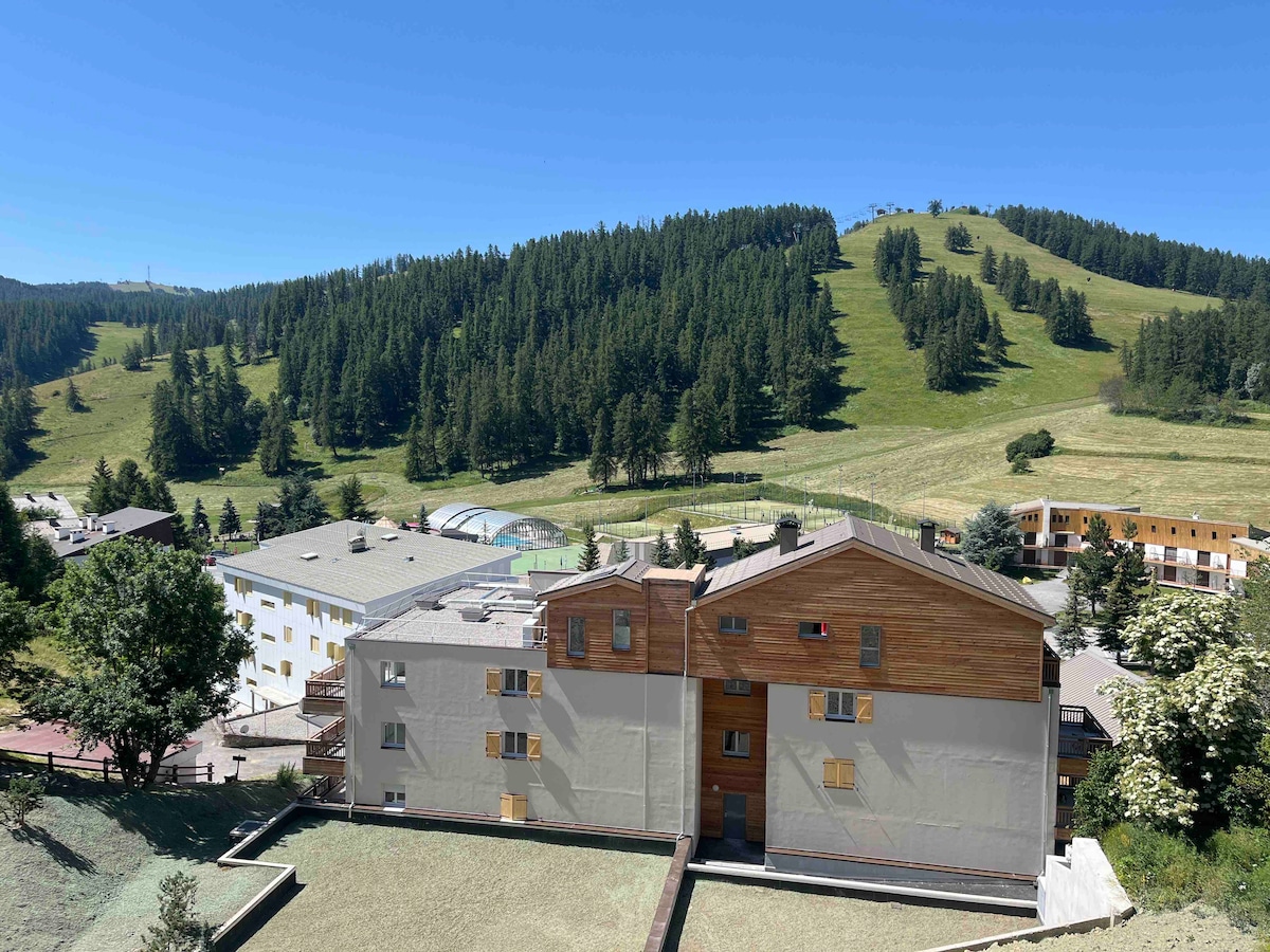 A view of the multifamily residence and surrounding landscape captures the natural beauty of lush green hills and tree-covered slopes. The building features a combination of natural wood and light-colored materials, set against a clear blue sky.