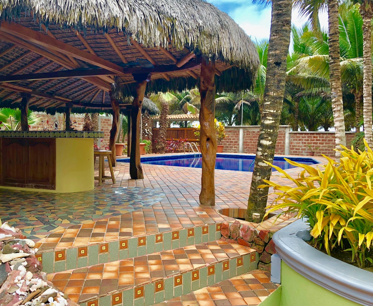 An outdoor area features a thatched-roof structure beside a sparkling pool, surrounded by lush greenery. The tiled floor displays a colorful pattern, and a bar area is visible, with a wooden stool positioned near it. Palm trees create a tropical ambiance in the background.