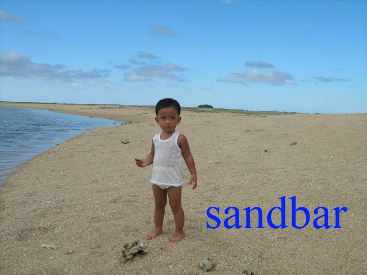 A sandy beach area is captured with gentle waves lapping at the shore. A child is positioned on the sand, surrounded by a wide expanse of beach. The sky above is clear, with a few clouds scattered, creating a serene coastal setting.