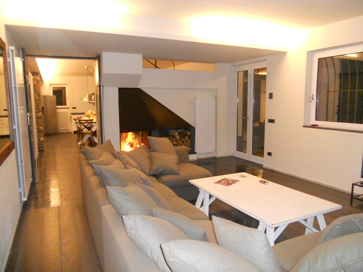 A spacious living area is furnished with a large, neutral-colored sectional sofa facing a modern bifacial fireplace. A light wooden coffee table sits in front, and large windows provide natural light. Open access to the kitchen and dining area enhances the flow of the space.