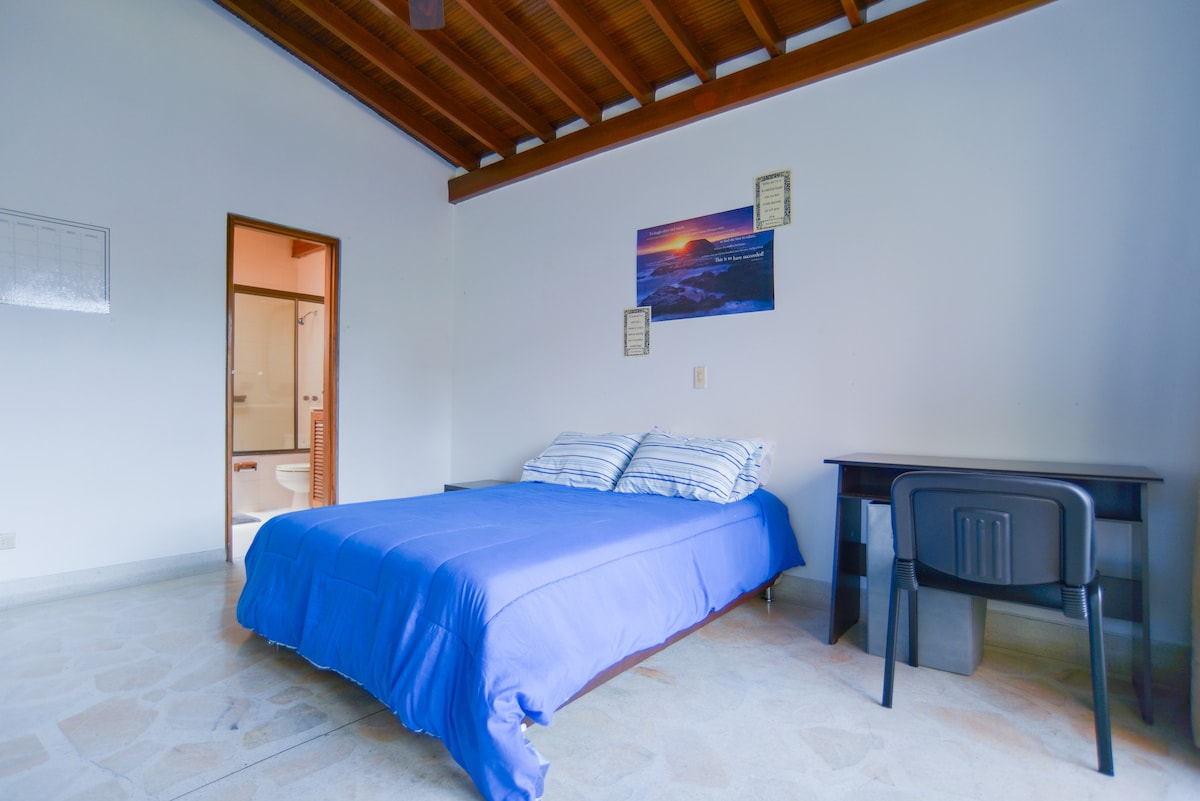 A well-lit bedroom features a spacious bed adorned with a blue coverlet and striped pillows. A functional desk with a chair is placed against one wall, while the entrance to an en suite bathroom is visible in the background.