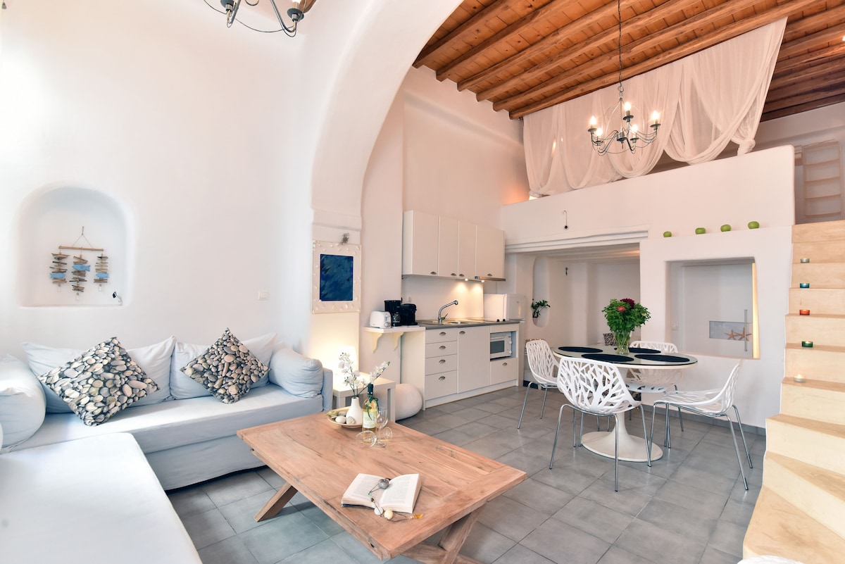 An airy living space showcases a light-colored couch adorned with patterned cushions, a wooden coffee table, and a dining area with white chairs. Modern kitchen fixtures are integrated within white cabinetry, while wooden beams and soft lighting contribute to a cozy ambiance.
