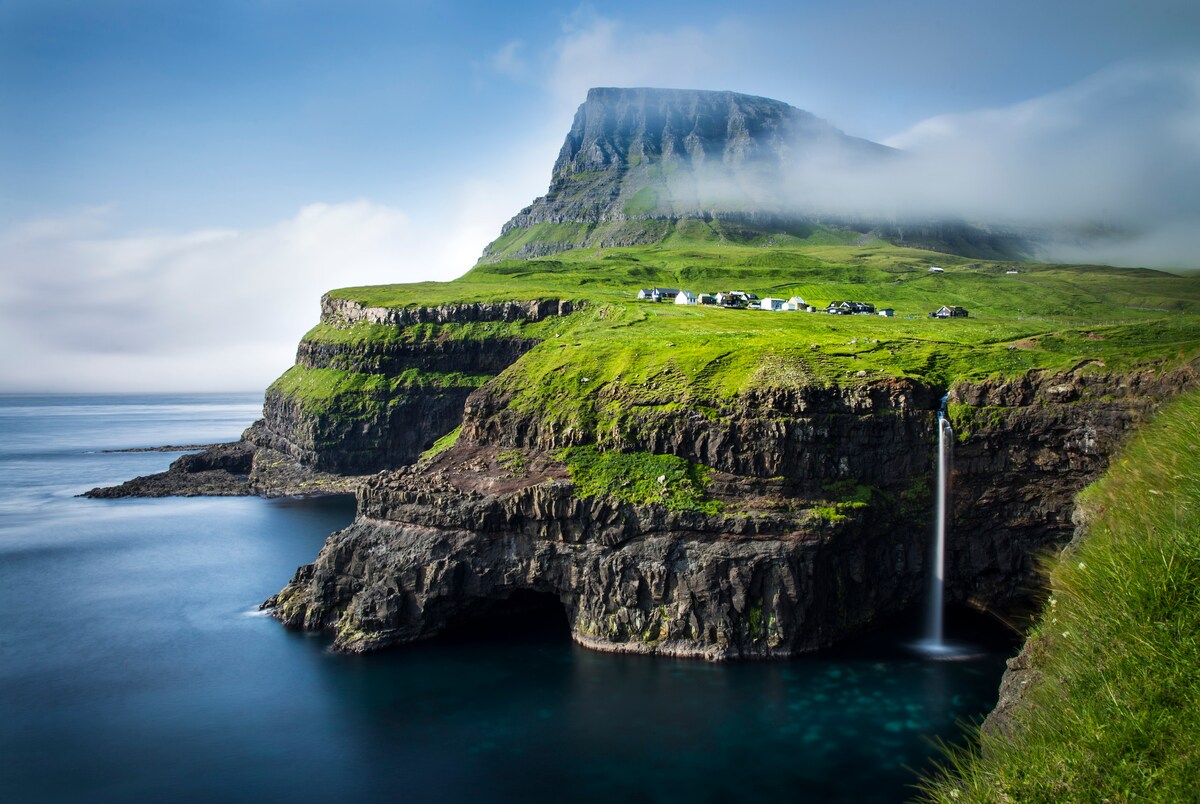 A dramatic coastal landscape is depicted, featuring steep, green cliffs meeting the calm ocean. A waterfall cascades into the water below, while a cluster of white buildings is visible atop the cliffs, surrounded by lush vegetation and misty mountains in the background.