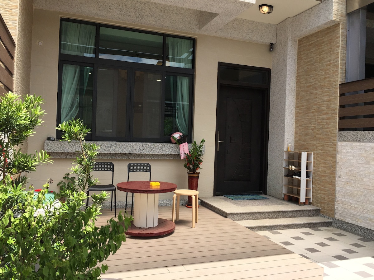 A welcoming outdoor area is featured, showcasing a wooden deck with a round table surrounded by four chairs. A small potted plant adds a touch of greenery, while the entrance to the room is framed by large windows adorned with sheer curtains.