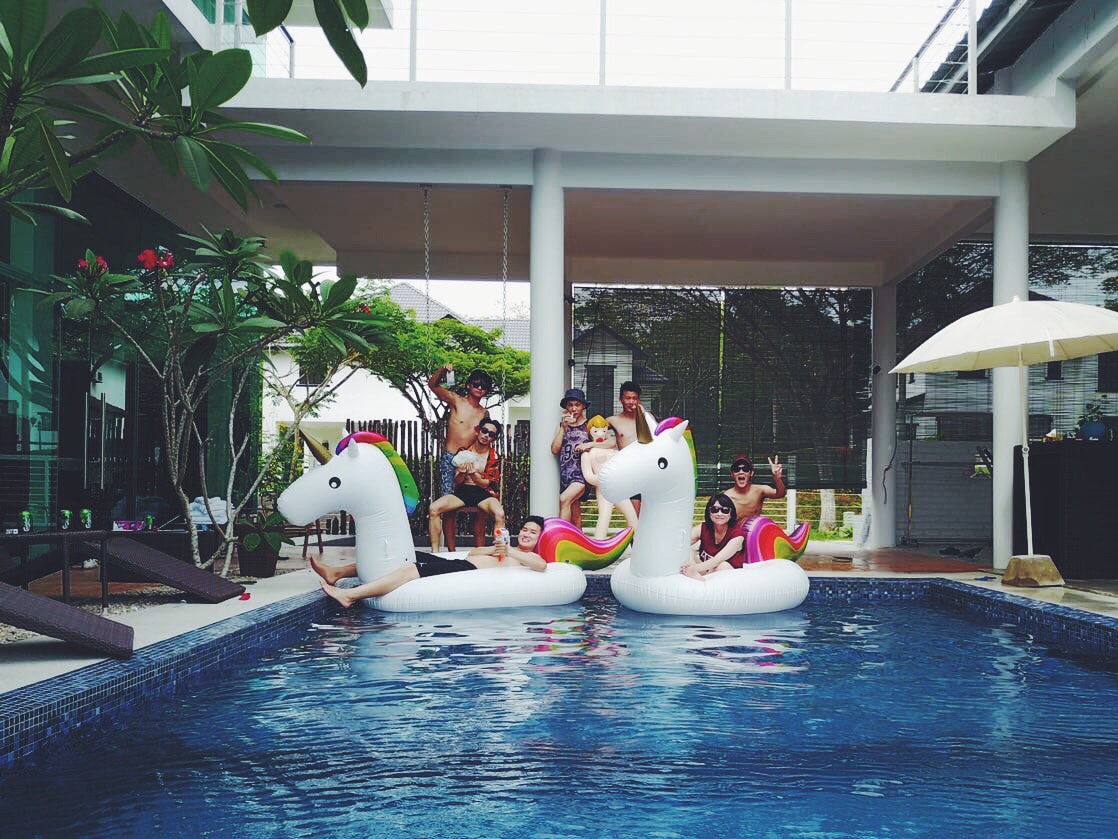 A private swimming pool features two unicorn-shaped floaties, occupied by guests enjoying the sun. Lush greenery surrounds the pool area, while sun loungers are set against a modern villa backdrop, enhancing the inviting atmosphere for relaxation.