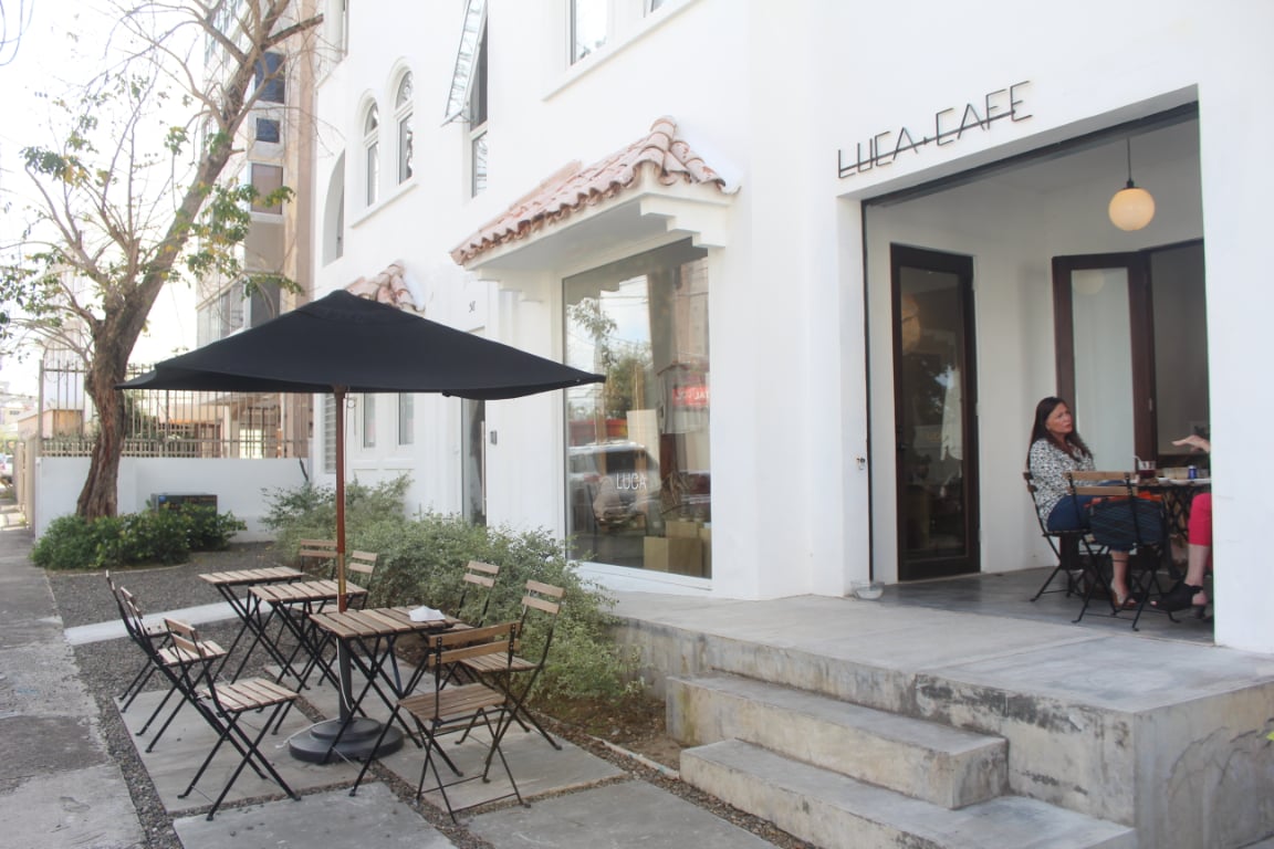 An outdoor café is featured with sleek, minimalist furnishings in a tranquil setting. A large black umbrella provides shade over several wooden tables and chairs on the patio, complemented by modern white architecture, showcasing arched windows and decorative tile accents.