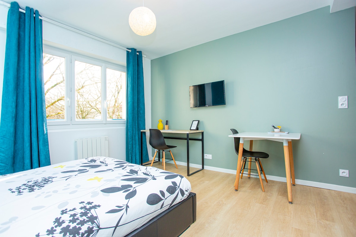 The room features a spacious bed adorned with floral bedding, complemented by bright teal curtains framing the windows. A wall-mounted television is visible, along with a desk and a small table, creating a functional and welcoming workspace.