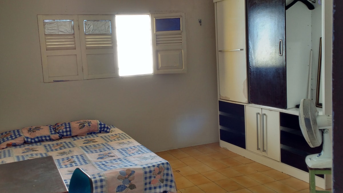 A well-lit room features a spacious bed with a patterned blanket and a flat surface table. A wardrobe with open shelving is present, and a fan is positioned nearby. Natural light enters through a window above the bed, illuminating the tiled floor.