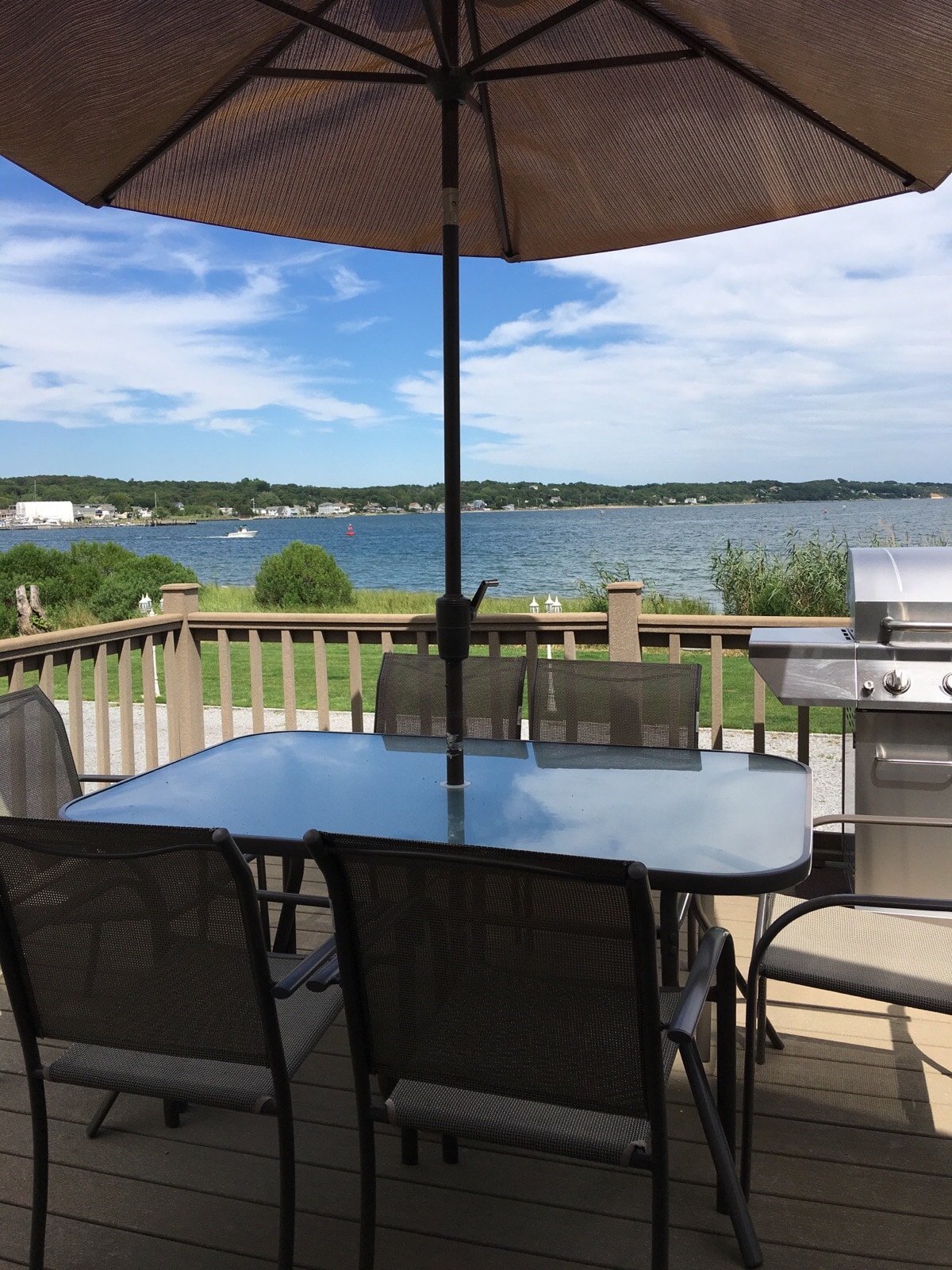 An outdoor dining area is visible, featuring a glass-top table surrounded by six chairs beneath a large umbrella. A grill sits nearby, with a view of the water and lush greenery in the background, creating a relaxing setting for meals and gatherings.