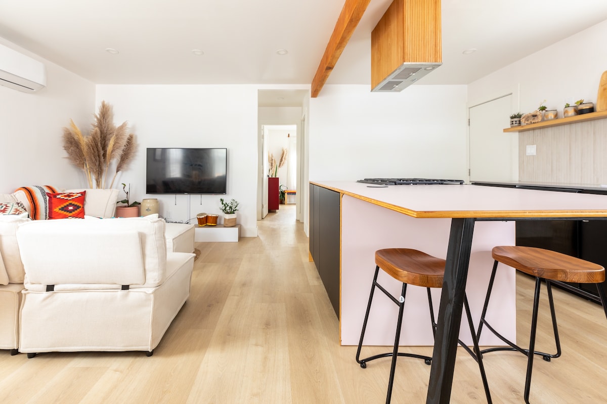 A modern living area is captured, featuring a plush white sectional sofa adorned with decorative pillows. A sleek television is mounted on the wall, while a kitchen island with wooden stools occupies the space opposite. Light wood flooring and minimalist decor contribute to an open, airy atmosphere.
