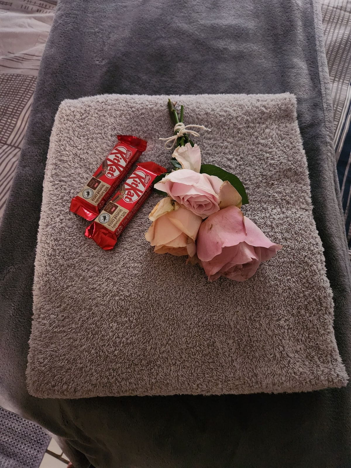 A soft grey towel is neatly folded, accompanied by two Kit Kat bars and a small bouquet of light pink and cream roses. The arrangement suggests a thoughtful touch for guests, creating a welcoming presentation.