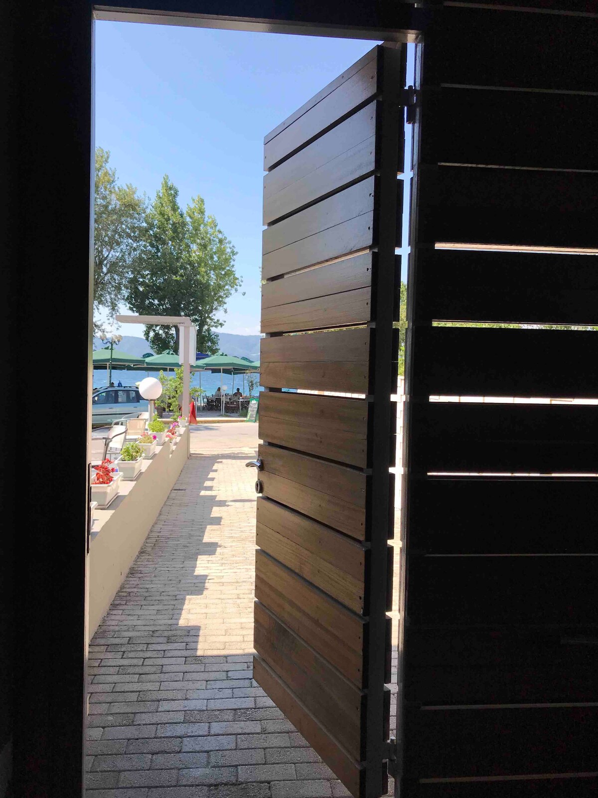 A partially open wooden door reveals a sunlit pathway lined with greenery, leading toward a scenic view of the water. Bright blue skies above enhance the inviting outdoor ambiance, while the walkway is paved with laid bricks, offering a sense of journey ahead.