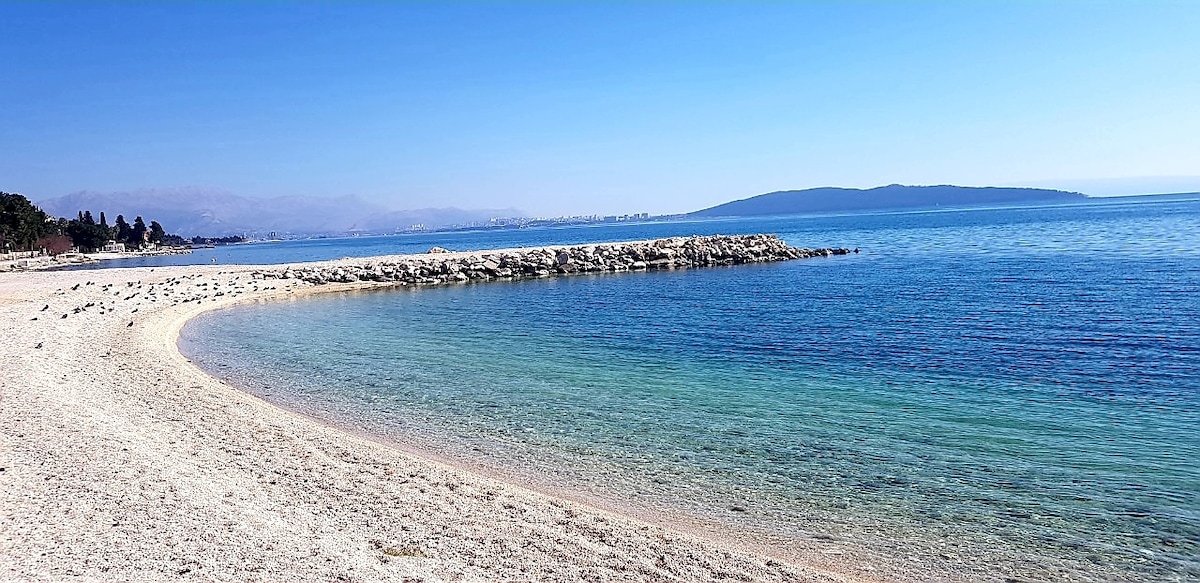 A serene beach scene is captured, showcasing a gentle curve of the sandy shoreline meeting calm, clear waters. Pebbles are scattered along the water's edge, while a distant island is visible against a clear blue sky, enhancing the sense of tranquility.