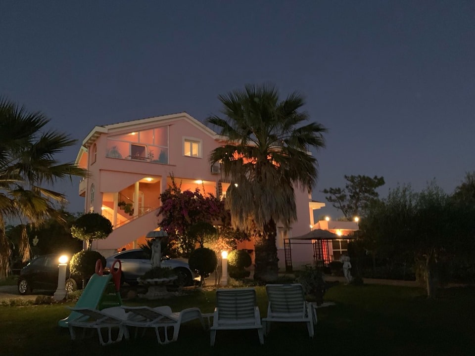 A charming multi-level building is illuminated at night, with soft lighting highlighting the palm trees in the foreground. The space features outdoor seating and a grassy lawn, creating a welcoming atmosphere for relaxation and enjoyment.