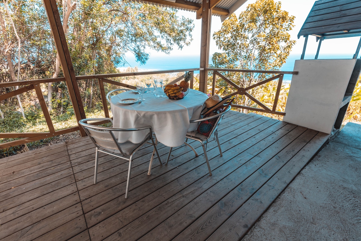 An outdoor dining area is presented with a round table covered by a white tablecloth, surrounded by four metal and wicker chairs. The deck is surrounded by wooden railing, offering views of the distant ocean visible through lush greenery.