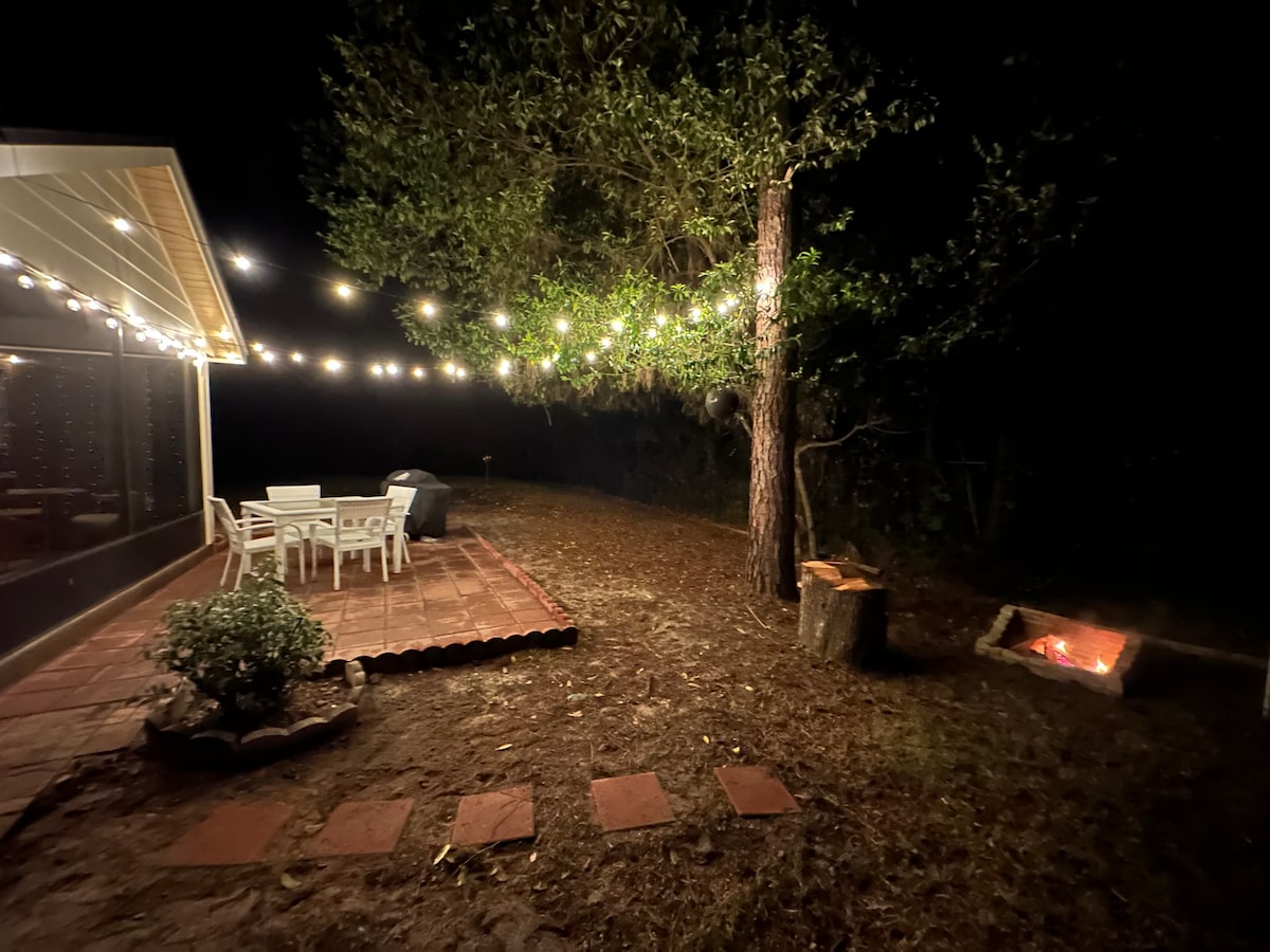 A tranquil outdoor space is illuminated by string lights, enhancing the inviting atmosphere. A dining table with chairs is situated beside a barbecue grill. A fire pit is visible in the corner, surrounded by a wooded area, providing a serene setting for evening gatherings.