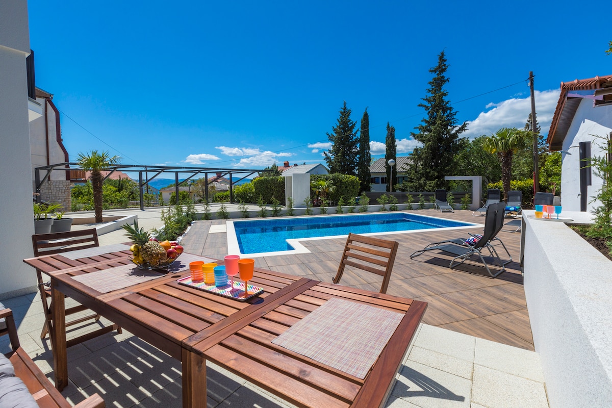 A covered terrace is shown with wooden dining furniture, including a table and chairs. A vibrant outdoor area features a swimming pool surrounded by sun loungers and landscaped greenery. The clear blue sky complements the relaxing atmosphere of the space.