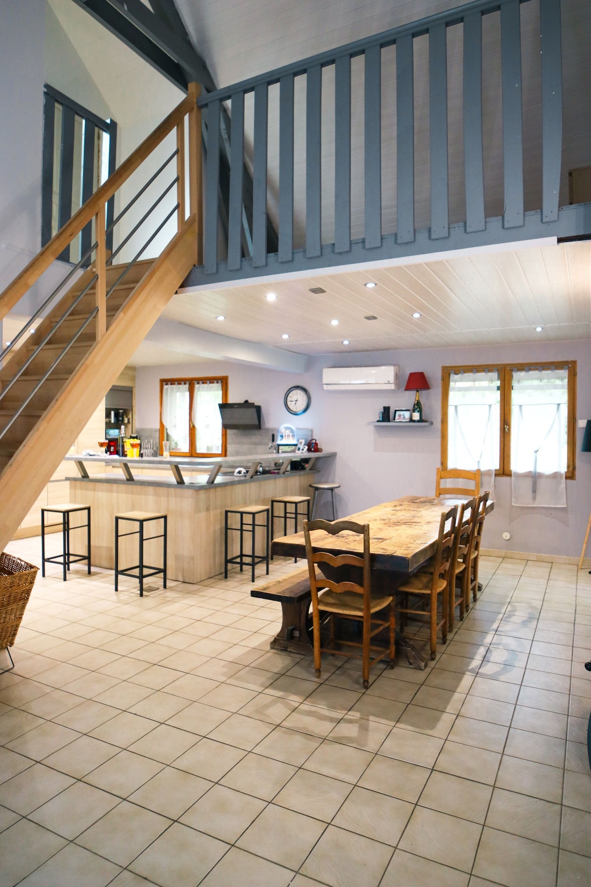 The spacious dining area features a large wooden table surrounded by several chairs, offering ample seating. A breakfast bar with stools is visible in the background. The room is illuminated by recessed ceiling lights, and large windows allow natural light to fill the space.
