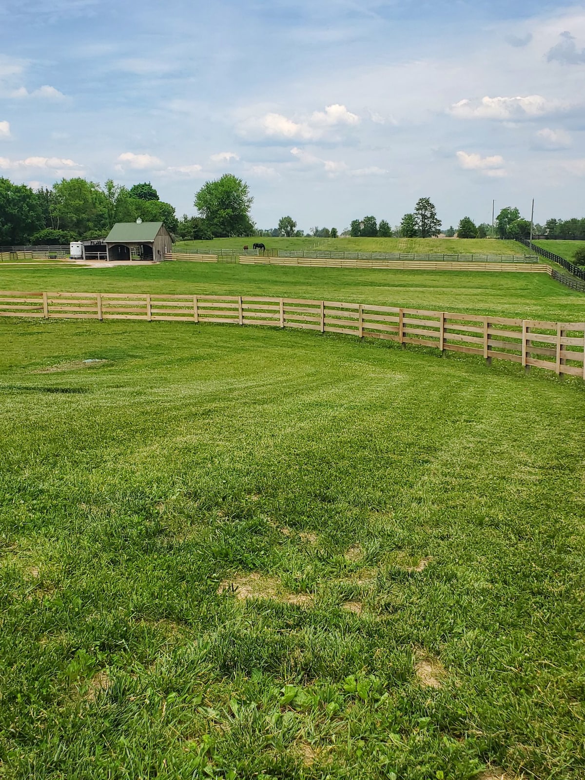 A lush, well-maintained green pasture is outlined by a wooden fence. In the background, a barn with a green roof is visible among the trees, with additional rolling fields beyond. The sky is slightly cloudy, contributing to a serene atmosphere.