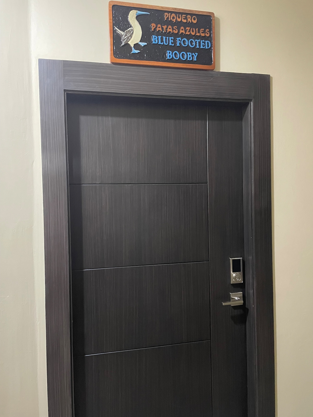 The entrance door of the suite features a modern design with a sleek, dark finish. A decorative sign labeled 'Piquero Patasazules Blue Footed Booby' hangs above the door, adding a unique touch. The door includes a digital lock for secure access.