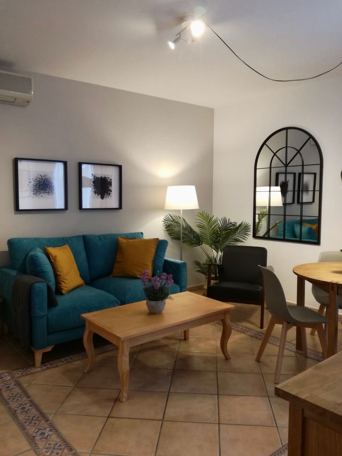 A cozy living area is depicted, featuring a teal sofa adorned with two yellow cushions. An elegant mirror reflects the soft glow from a nearby lamp, while a small wooden coffee table is centered among potted plants. Light beige tiles and neutral walls enhance the inviting space.