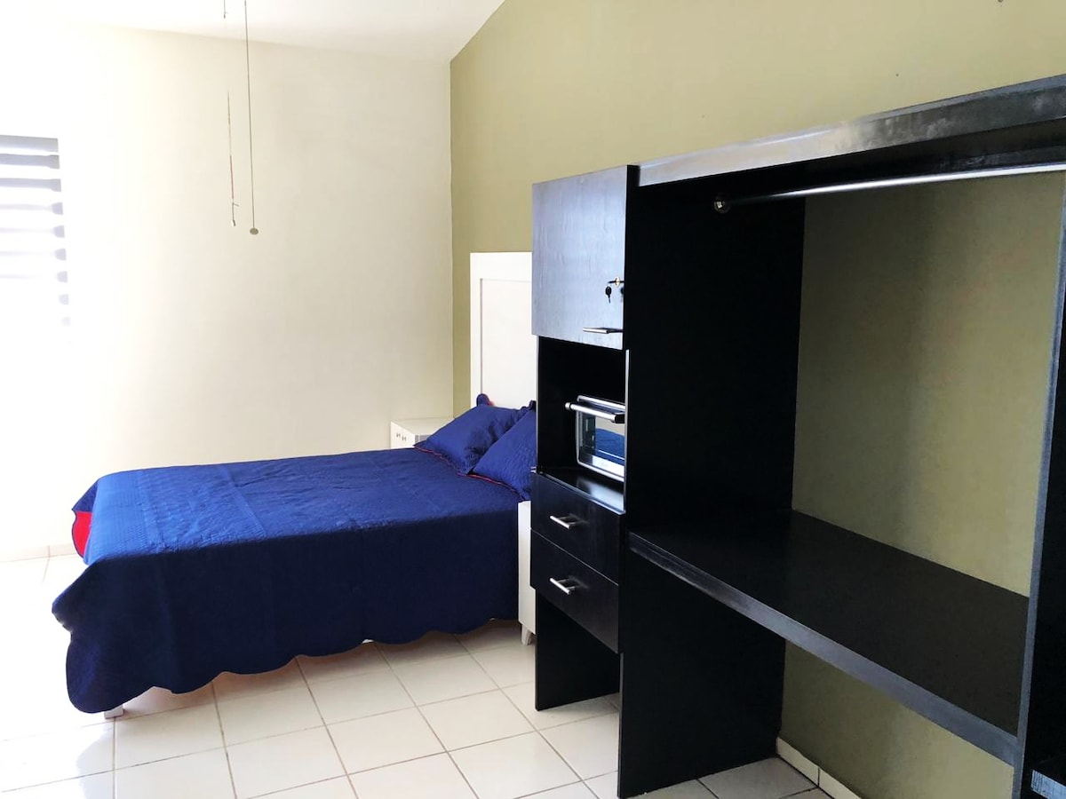A simple sleeping area is observed with a single bed draped in a blue coverlet. A contrasting dark furniture unit with drawers and a workspace is present. Natural light enters through a window, enhancing the room's bright ambience.