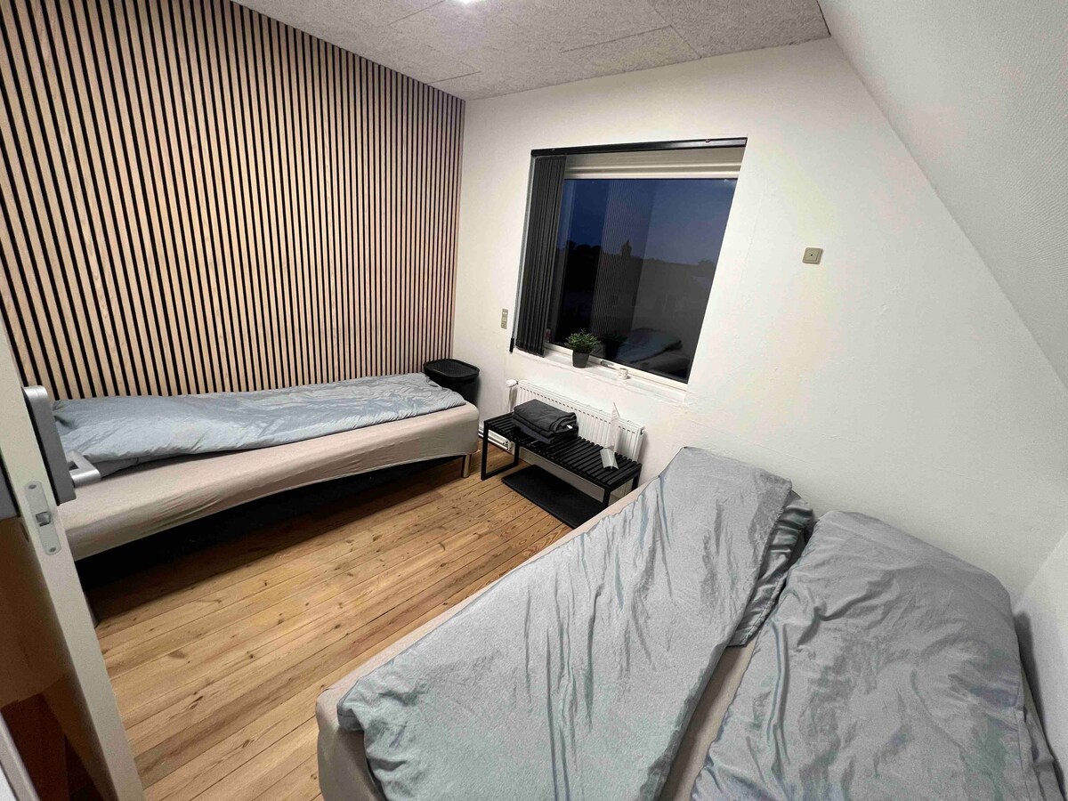 A room features two spacious beds with light-coloured bedding, arranged parallel to each other. Natural wood flooring adds warmth, while a large window offers views of the surroundings. A small black shelf beside the window holds essential items, enhancing the room's functionality.
