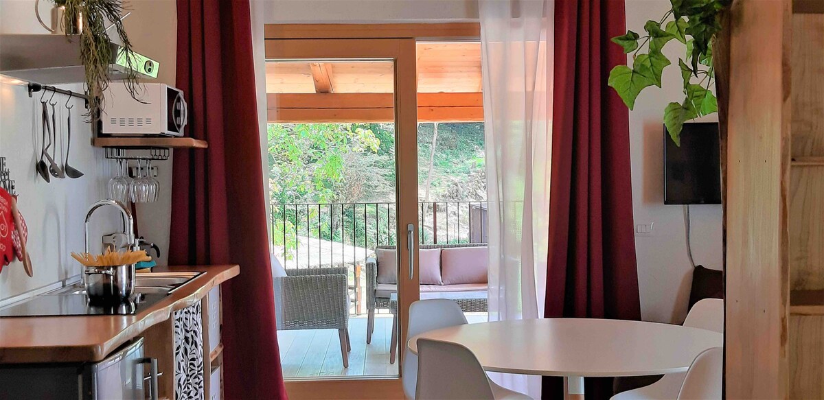 A bright and inviting kitchen area is visible, featuring a round dining table and white chairs. Large glass doors open to a covered outdoor space with comfortable seating, surrounded by greenery. Soft curtains frame the view, enhancing the connection to the outdoor environment.