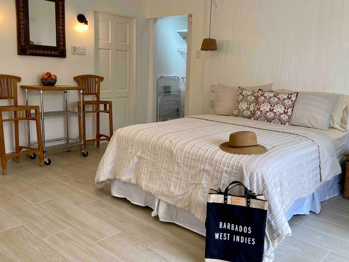 A spacious studio apartment features a large bed with neutral linens and decorative pillows. A small dining area with two wooden stools is positioned next to the bed. A straw hat and a canvas bag are placed on the floor, adding to the inviting ambiance.