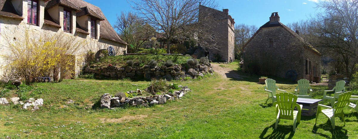A spacious garden area features well-maintained green grass, surrounded by traditional stone buildings. Colorful seating is arranged in a circular layout, providing a relaxed space for guests. A small stone path leads through the garden, inviting exploration of the natural surroundings.